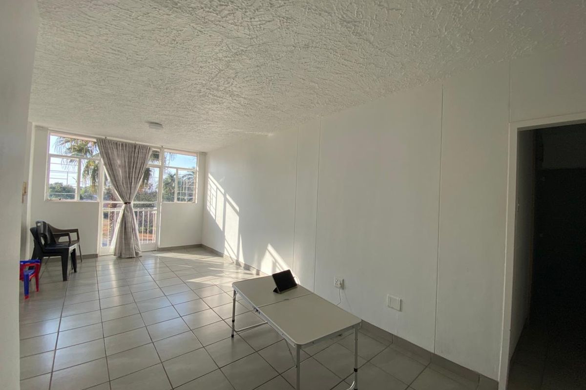 A bright, open-plan living area featuring neutral tiled flooring, a textured ceiling, and large windows providing ample natural light with access to a balcony.