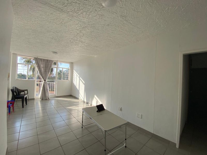 A bright, open-plan living area featuring neutral tiled flooring, a textured ceiling, and large windows providing ample natural light with access to a balcony.