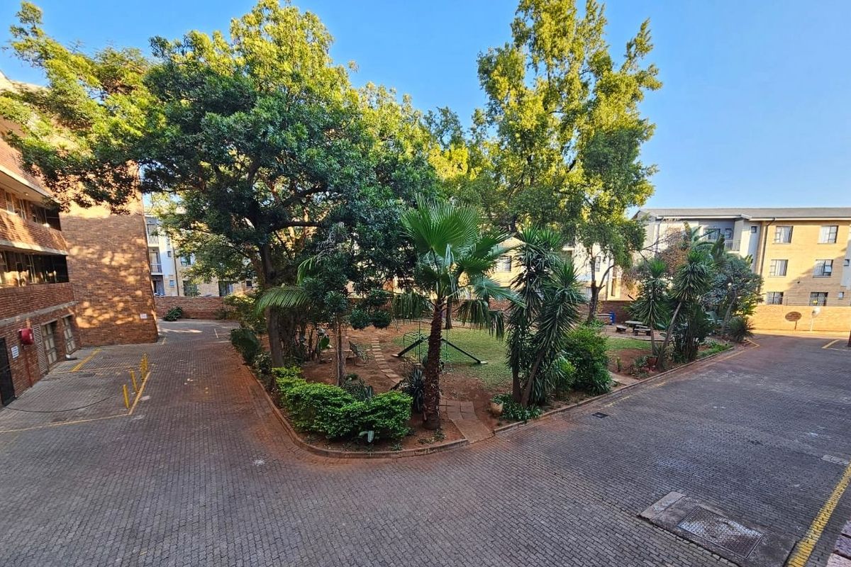A view of the apartment complex's communal garden area, featuring mature trees, landscaped greenery, and paved driveway access.