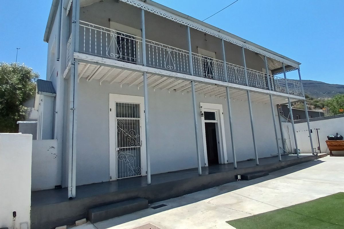A two-story rear exterior view featuring a covered veranda with wrought iron balustrades, concrete patio area, and artificial lawn.