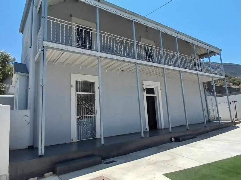 A two-story rear exterior view featuring a covered veranda with wrought iron balustrades, concrete patio area, and artificial lawn.