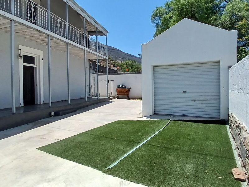 Rear exterior view featuring a detached garage, concrete driveway, artificial lawn, and a two-story building with a balcony.