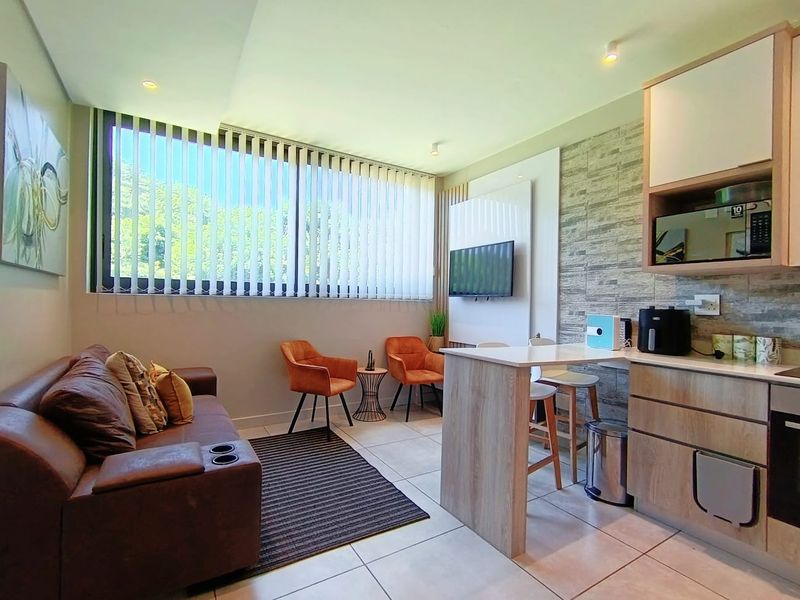 Modern open-plan living area featuring a compact kitchen with breakfast bar, stone-look feature wall, and large windows with vertical blinds.