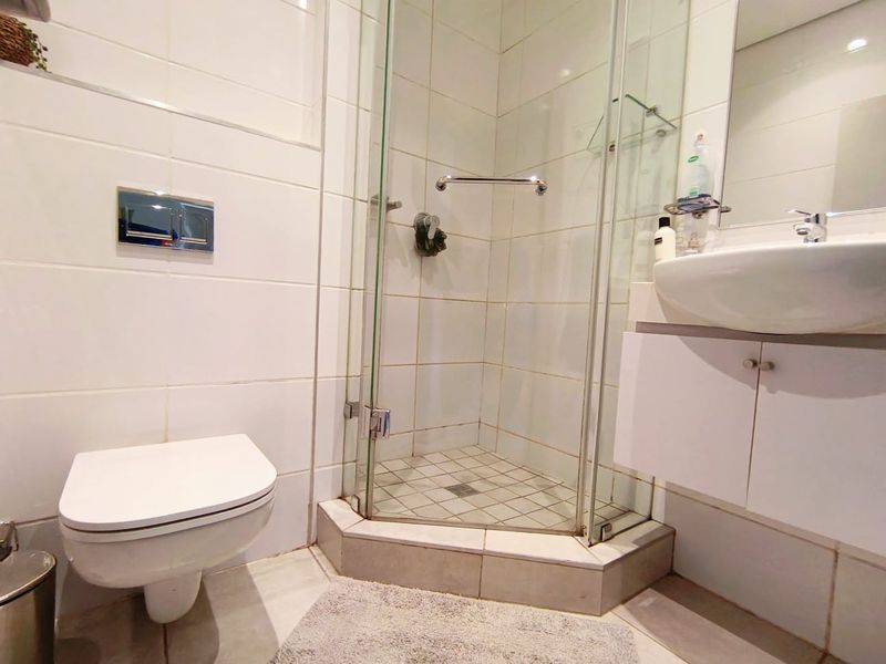 Compact bathroom featuring a wall-mounted toilet, glass shower enclosure, and a vanity unit with a basin, finished with white wall tiles.