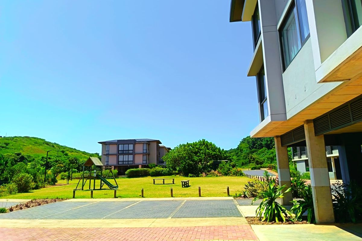A view of the apartment complex exterior featuring a paved parking area, landscaped communal garden, and modern building architecture.