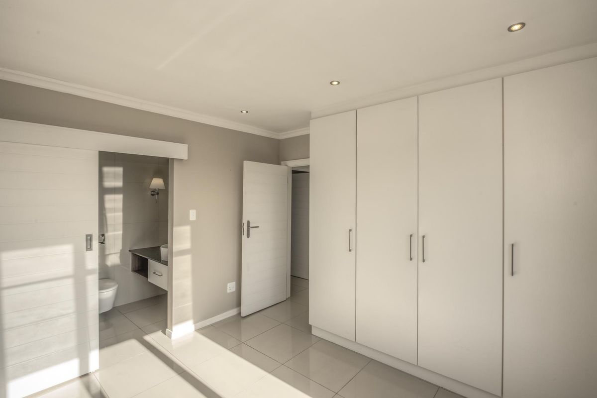A bright, modern bedroom featuring neutral tones, tiled flooring, ample built-in cupboard space, and direct access to an en-suite bathroom.