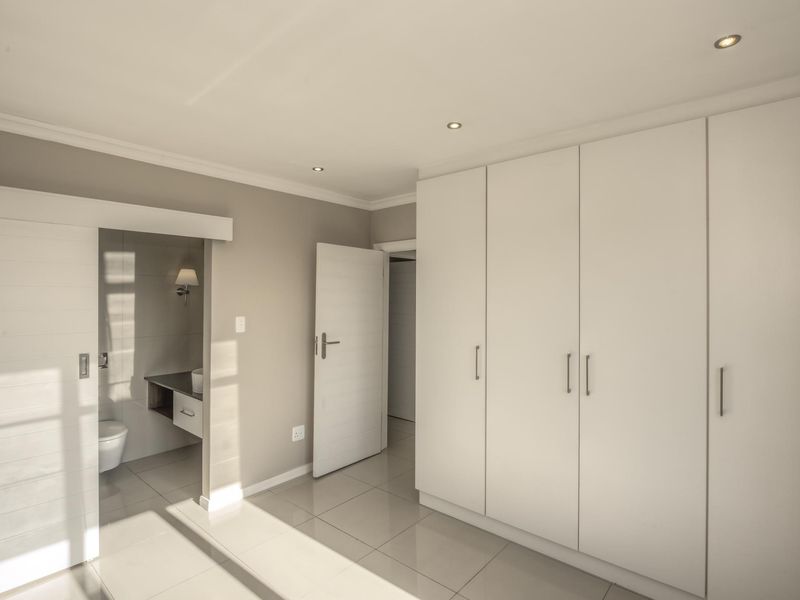 A bright, modern bedroom featuring neutral tones, tiled flooring, ample built-in cupboard space, and direct access to an en-suite bathroom.