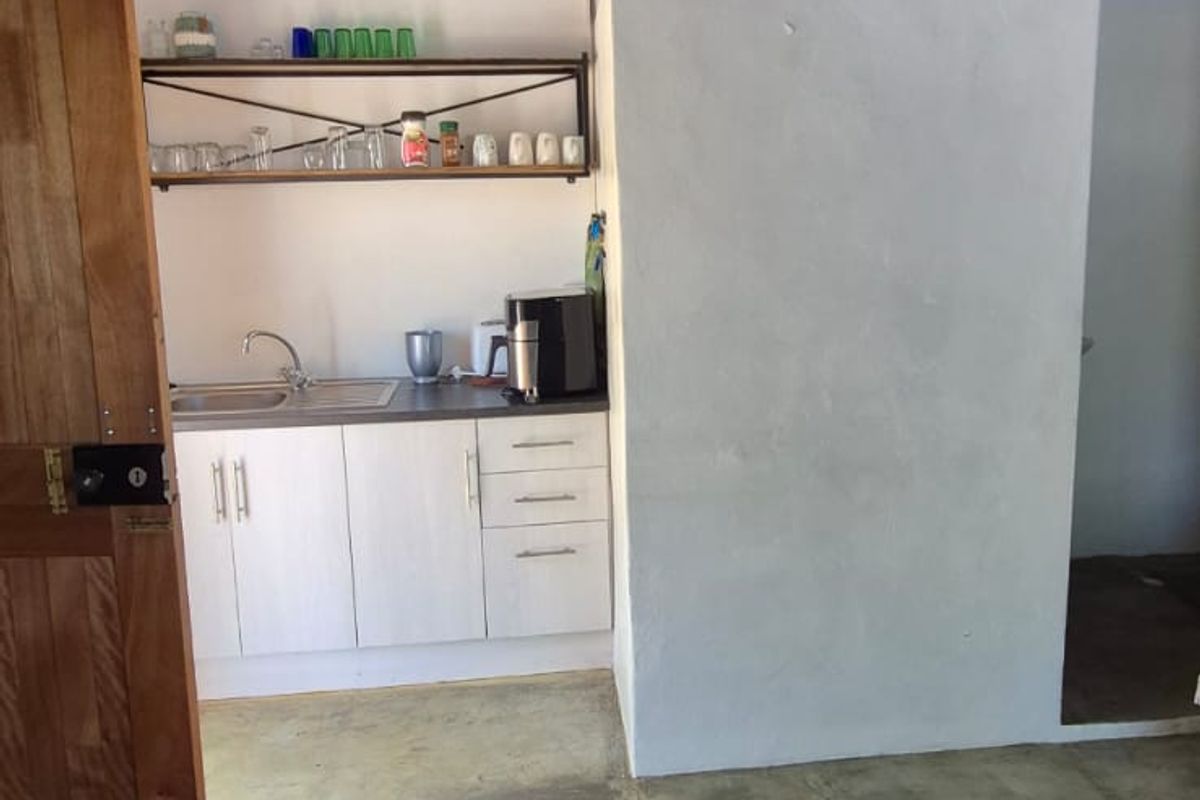 A compact kitchenette area featuring built-in cabinetry, a stainless steel sink, open shelving, and exposed timber ceiling beams with a concrete floor finish.