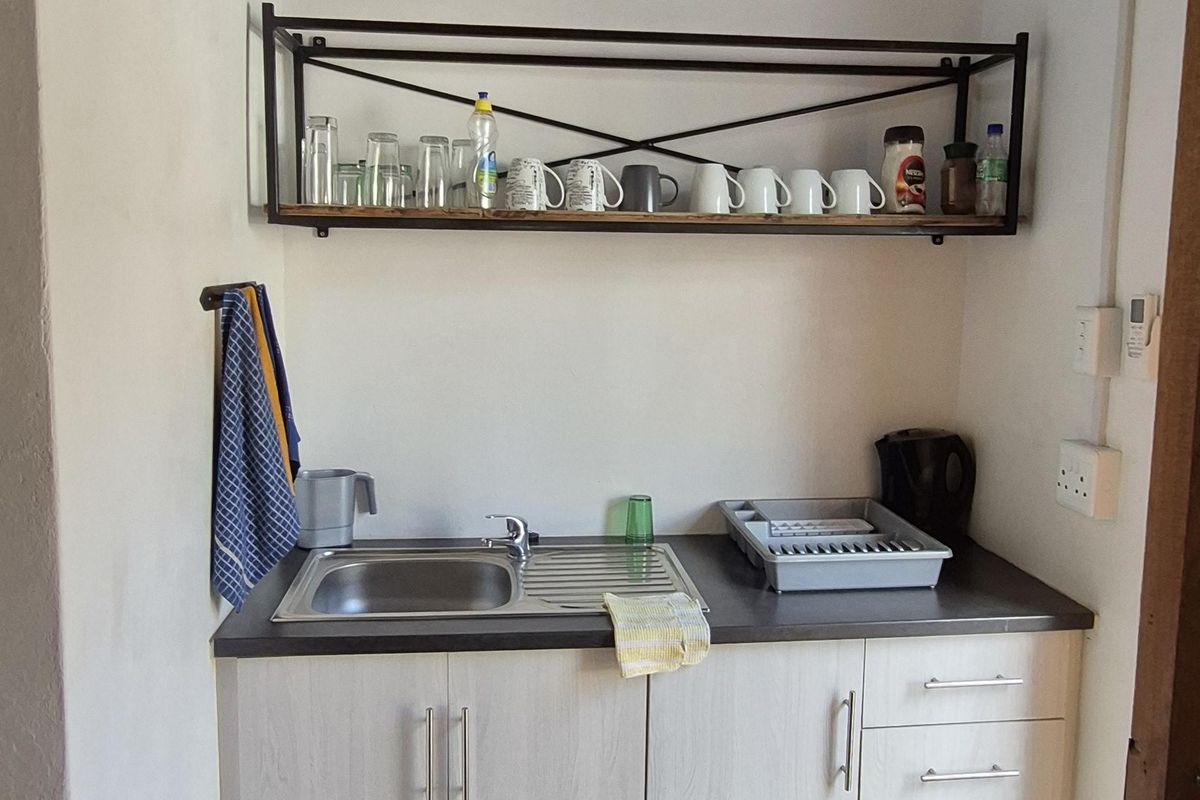 A compact scullery area featuring light-toned cabinetry, a stainless steel sink, and open metal shelving against a white wall.