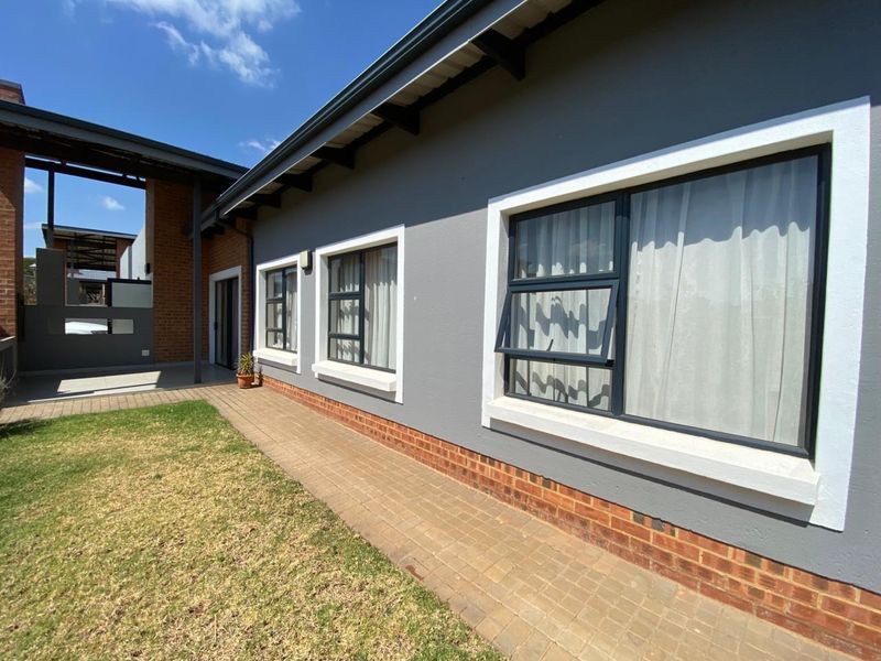 Rear exterior view of the house featuring a paved walkway, grey painted walls with brick detailing, and modern aluminium window frames.