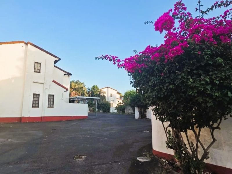 Exterior view of the townhouse complex featuring an asphalt driveway, white stucco facade with red trim, and a carport area.