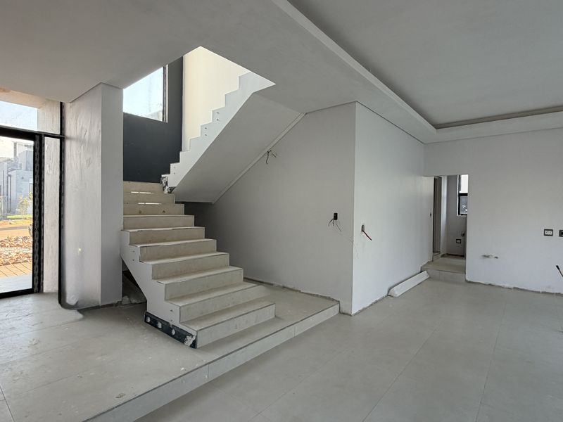 An open-plan living area under construction, featuring a modern staircase, tiled flooring, and recessed ceiling details, awaiting final finishes and electrical fixtures.