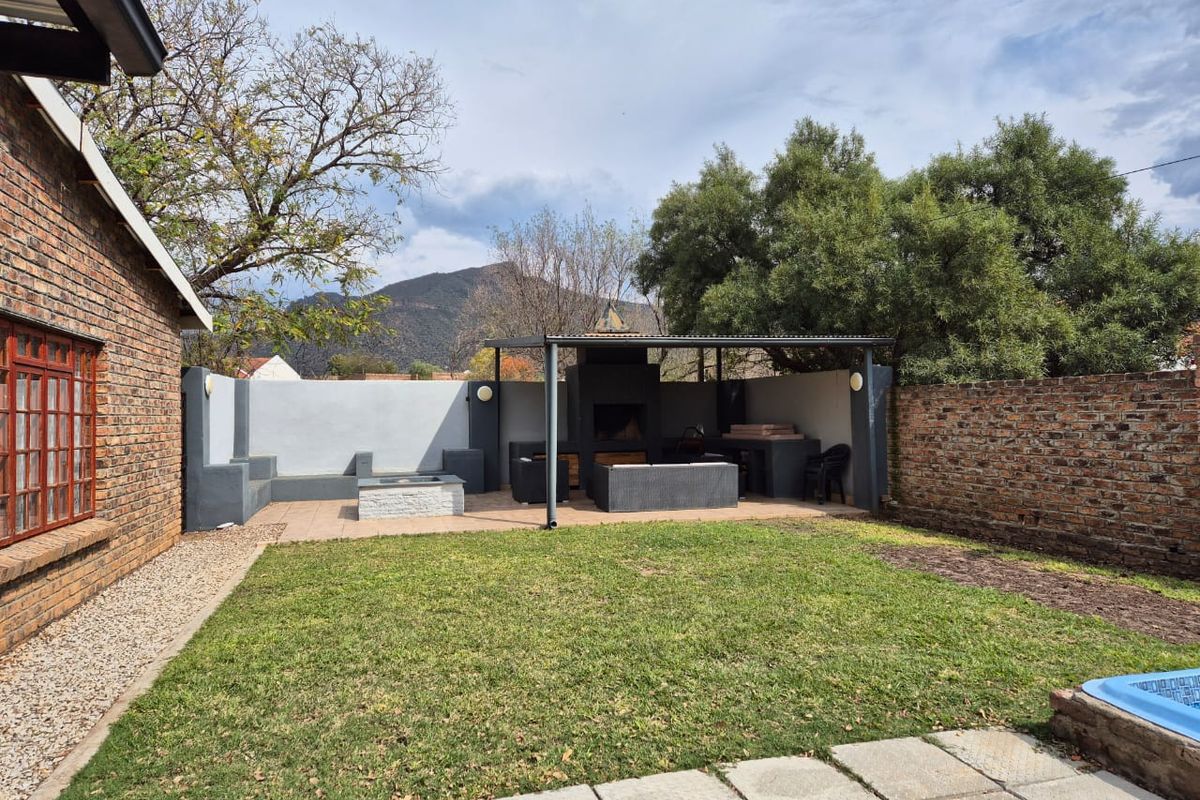 A well-maintained rear garden featuring a covered patio area with a built-in braai, brick boundary walls, and a lawn.