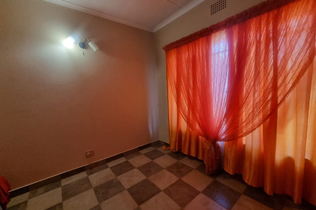 An empty bedroom featuring checkered tiled flooring, a wall-mounted light fixture, and large windows covered by orange curtains.