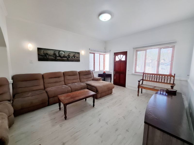 A bright living room featuring light-toned laminate flooring, wall sconces, and a classic timber front door with natural light through venetian blinds.