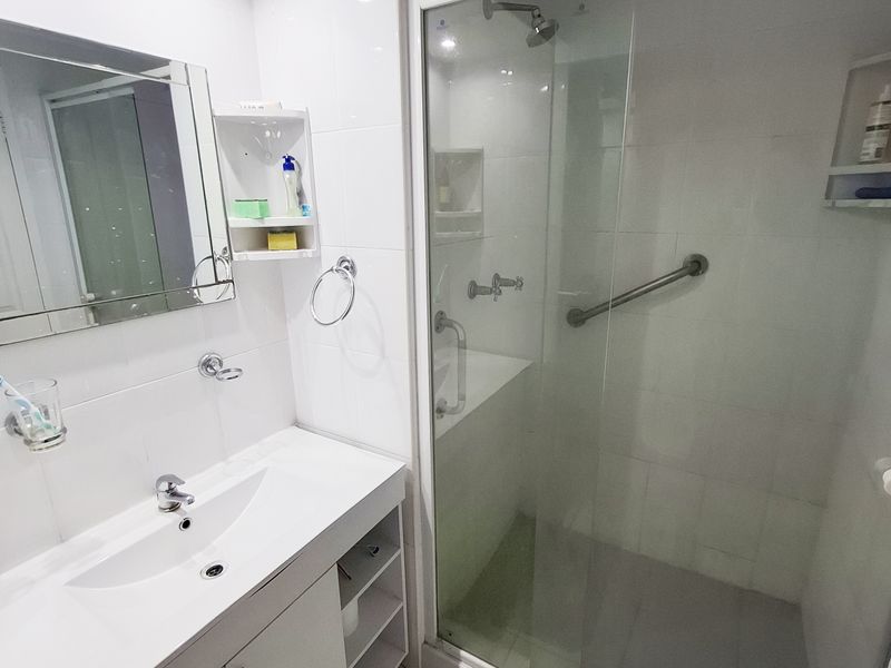 A clean, modern bathroom featuring a white vanity with integrated basin, a glass-enclosed shower with a safety grab rail, and a mirrored medicine cabinet.