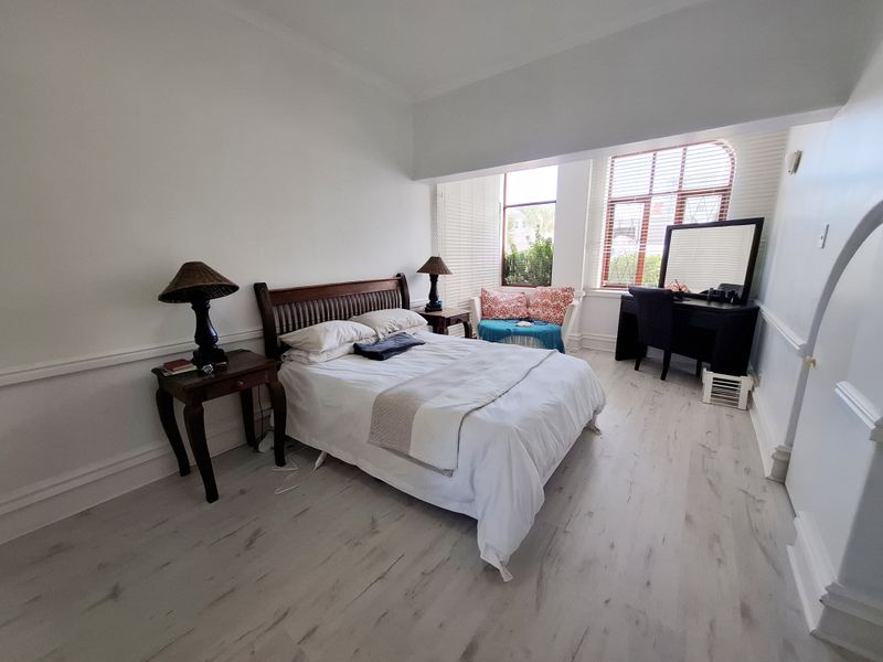 A bright and spacious bedroom featuring light-toned laminate flooring, classic wainscoting, and large windows with venetian blinds allowing for ample natural light.