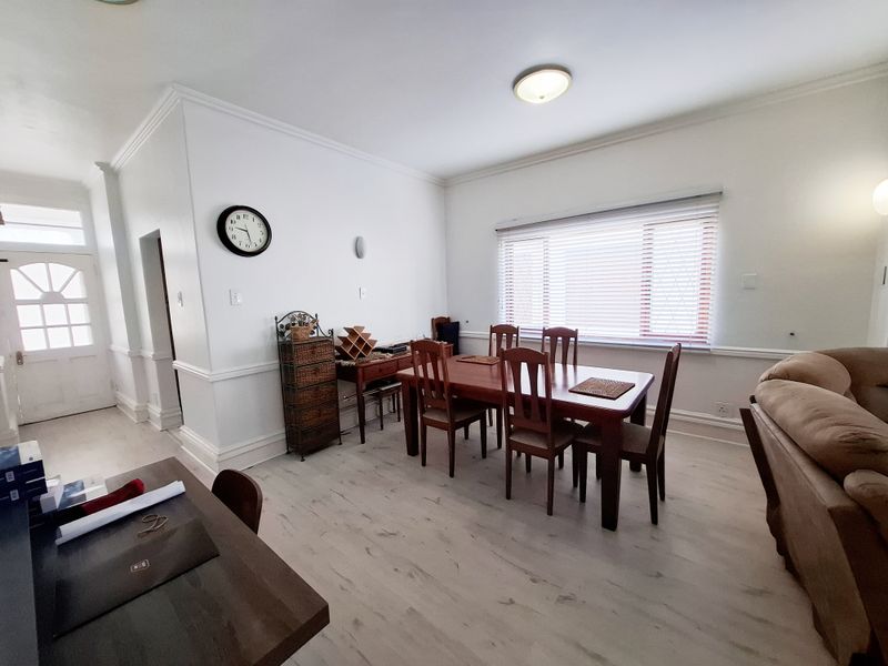 A bright dining area featuring light-toned laminate flooring, classic crown molding, chair rail detailing, and large windows fitted with venetian blinds.