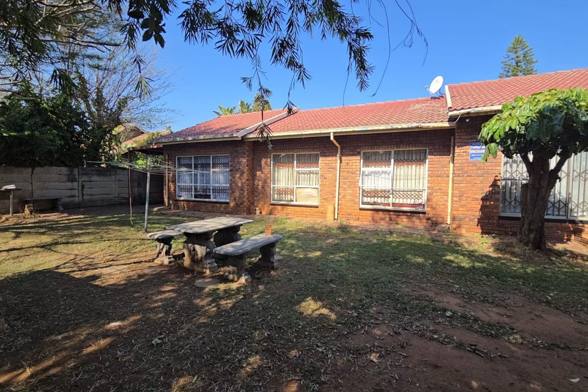 Rear view of a single-story brick house featuring a tiled roof, security bars on windows, and a spacious backyard with a boundary wall.