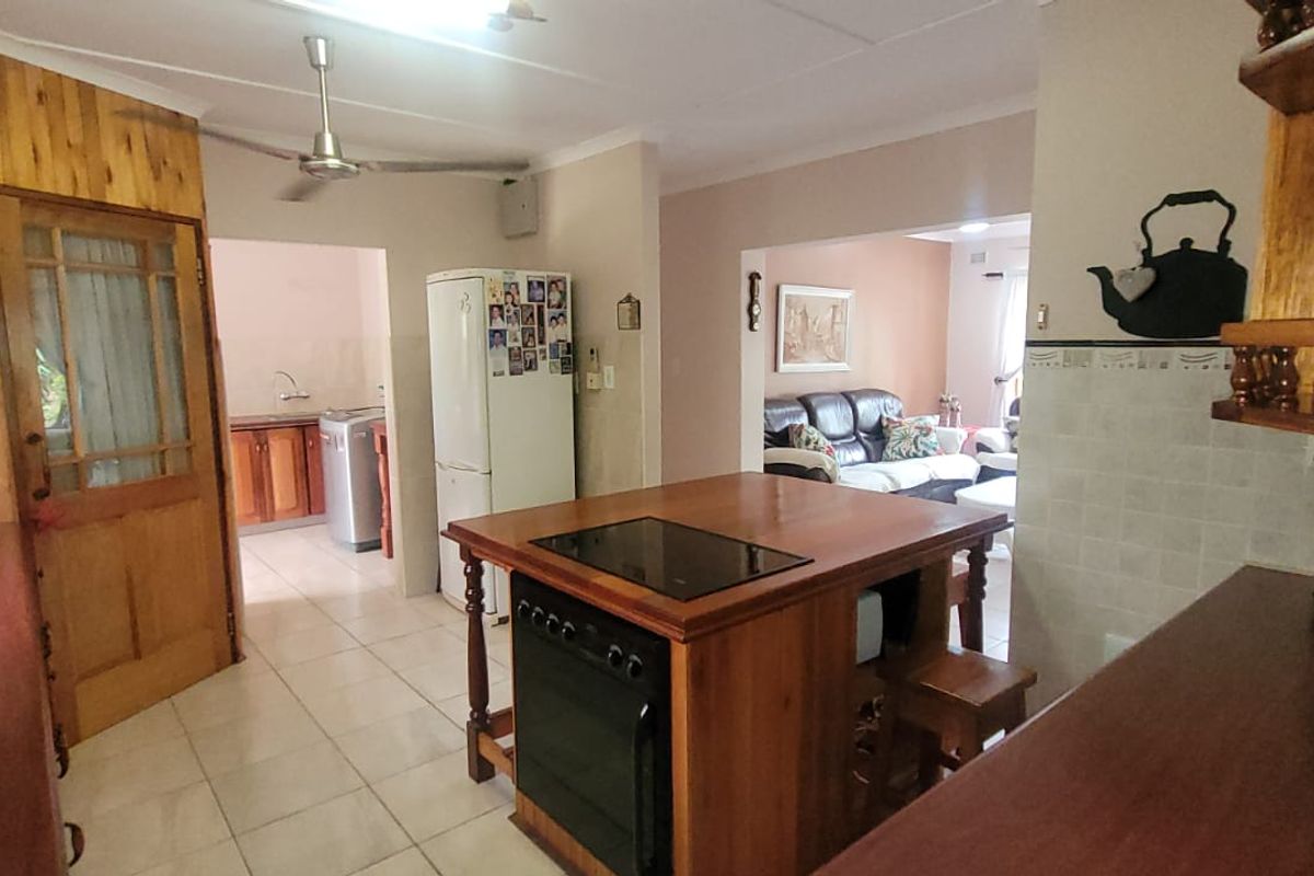 A functional kitchen featuring wooden cabinetry, a central island with a built-in oven and cooktop, and tiled flooring, connecting to an adjacent living area.