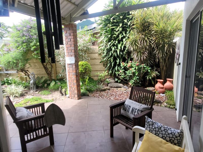 A covered outdoor patio area featuring tiled flooring, a brick support pillar, and direct access to a landscaped garden through sliding glass doors.