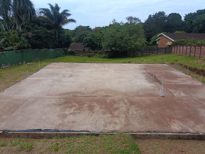 A vacant residential plot featuring a prepared concrete slab foundation with visible plumbing rough-ins, ready for new construction.