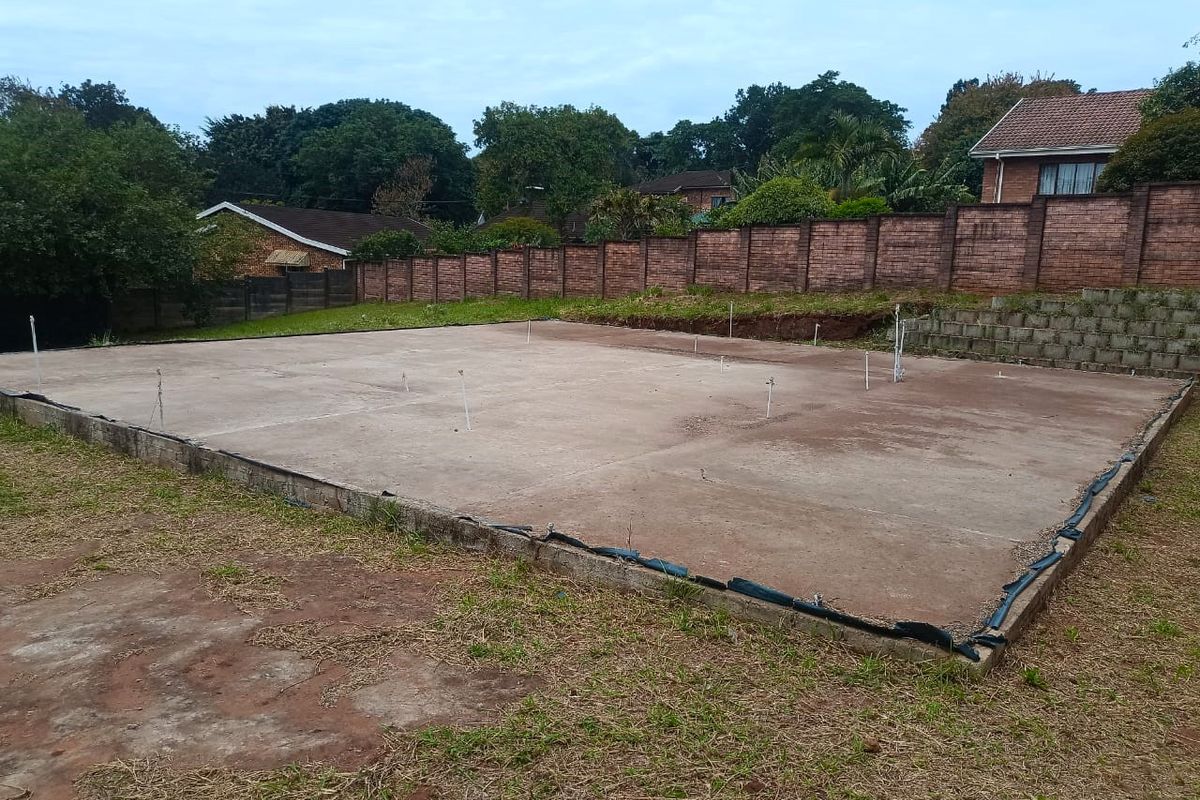 A vacant residential plot featuring a prepared concrete slab foundation with plumbing rough-ins, enclosed by a brick boundary wall and retaining wall.