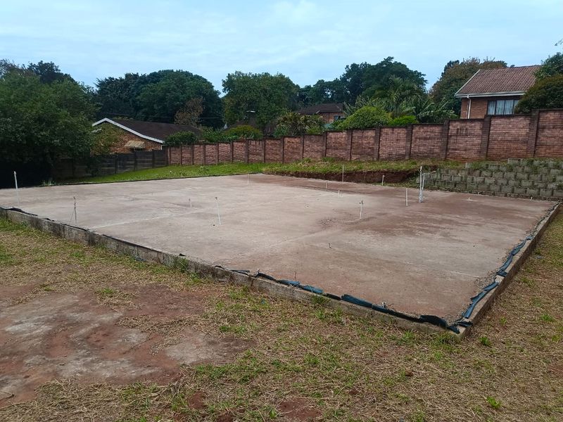 A vacant residential plot featuring a prepared concrete slab foundation with plumbing rough-ins, enclosed by a brick boundary wall and retaining wall.