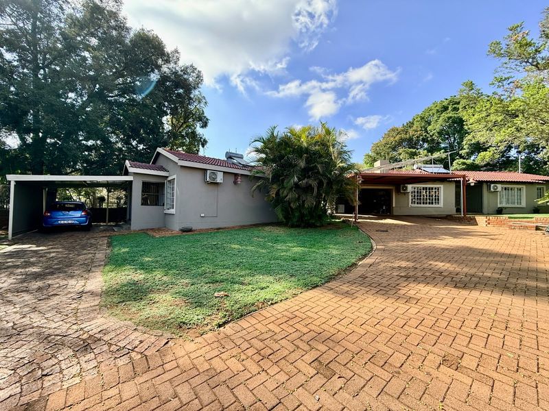 Front view of the property featuring a paved driveway, carport, landscaped lawn, and established trees under a clear sky.