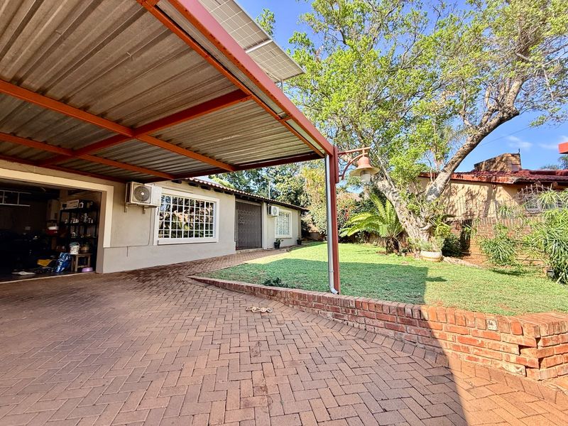 Rear exterior view featuring a paved driveway, covered carport with solar panels, brick retaining wall, and a landscaped garden area.