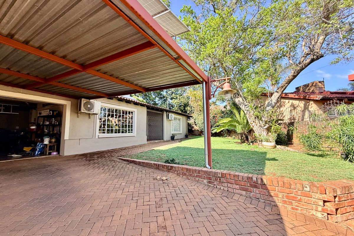 Rear exterior view featuring a paved driveway, covered carport with solar panels, brick retaining wall, and a landscaped garden area.