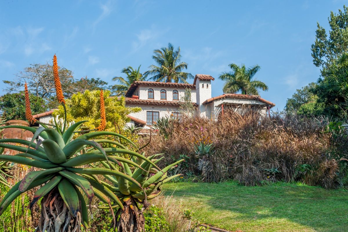 A view of the rear elevation of a Mediterranean-style house featuring a terracotta tiled roof, stucco walls, and arched windows set within a landscaped garden.