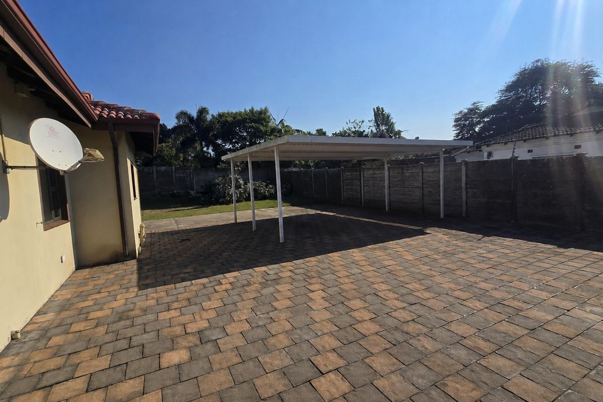 A spacious rear exterior area featuring a paved driveway, a functional carport structure, and secure precast concrete boundary walls.