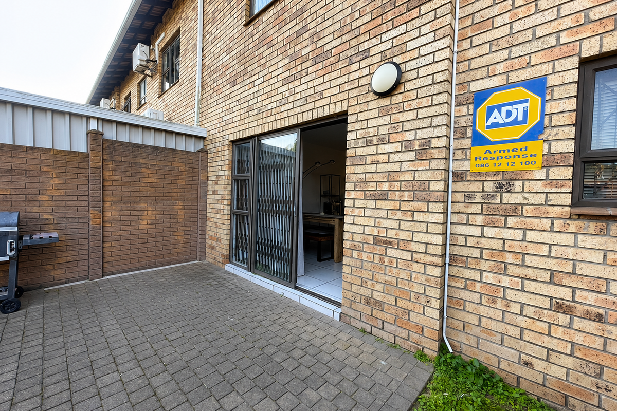 Rear exterior view of the townhouse featuring brick paving, a sliding door with security gate, and an external wall-mounted security sign.