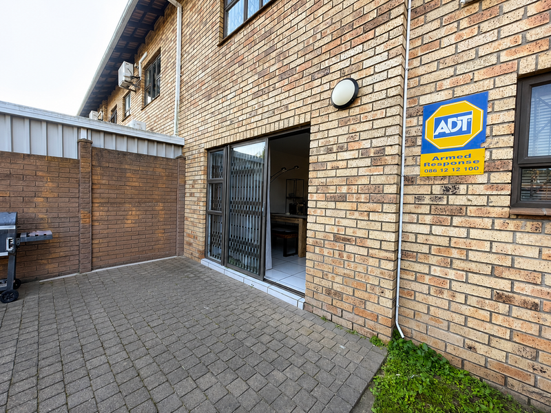 Rear exterior view of the townhouse featuring brick paving, a sliding door with security gate, and an external wall-mounted security sign.