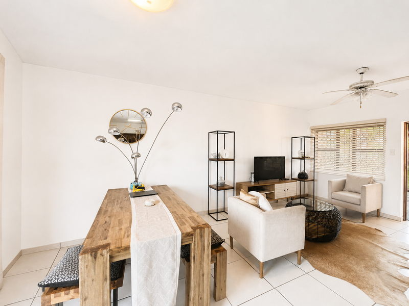 An open-plan living and dining area featuring neutral tiled flooring, a ceiling fan, and natural light through venetian blinds.