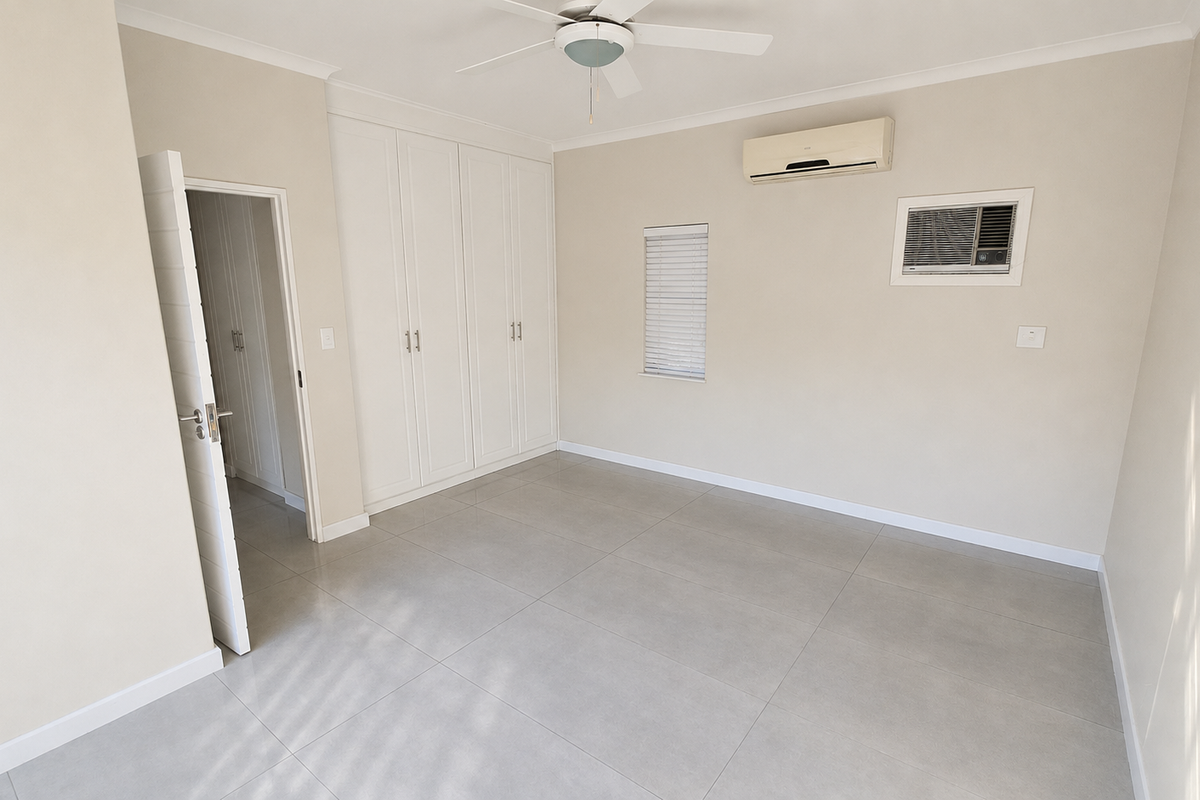 A spacious, empty bedroom featuring neutral tiled flooring, built-in cupboards, a ceiling fan, and dual air conditioning units for climate control.