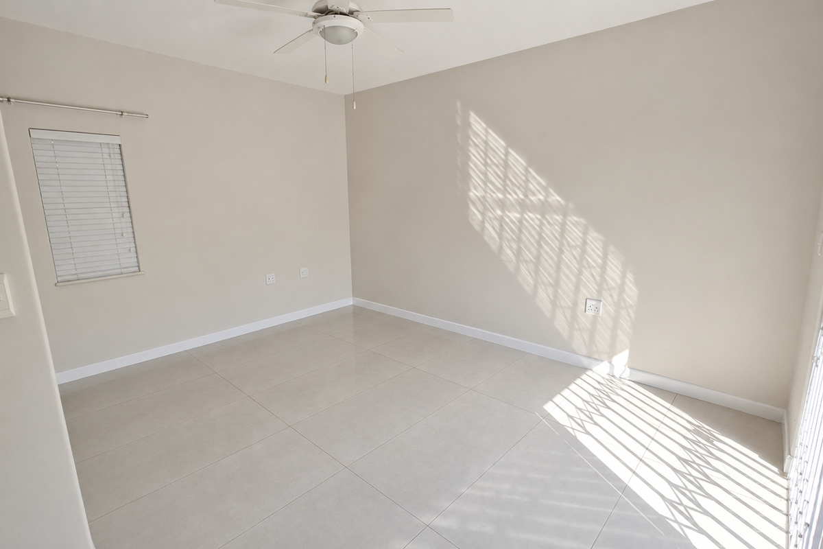 A bright, empty living area featuring neutral tiled flooring, a ceiling fan, and access to an outdoor space through a security gate.