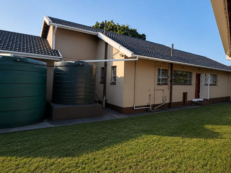 Rear view of the property featuring a lawn, concrete pathways, two large water storage tanks, and a built-in outdoor braai area.