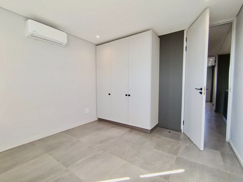 A bright, modern bedroom featuring neutral tiled flooring, built-in white cabinetry, and a wall-mounted air conditioning unit.