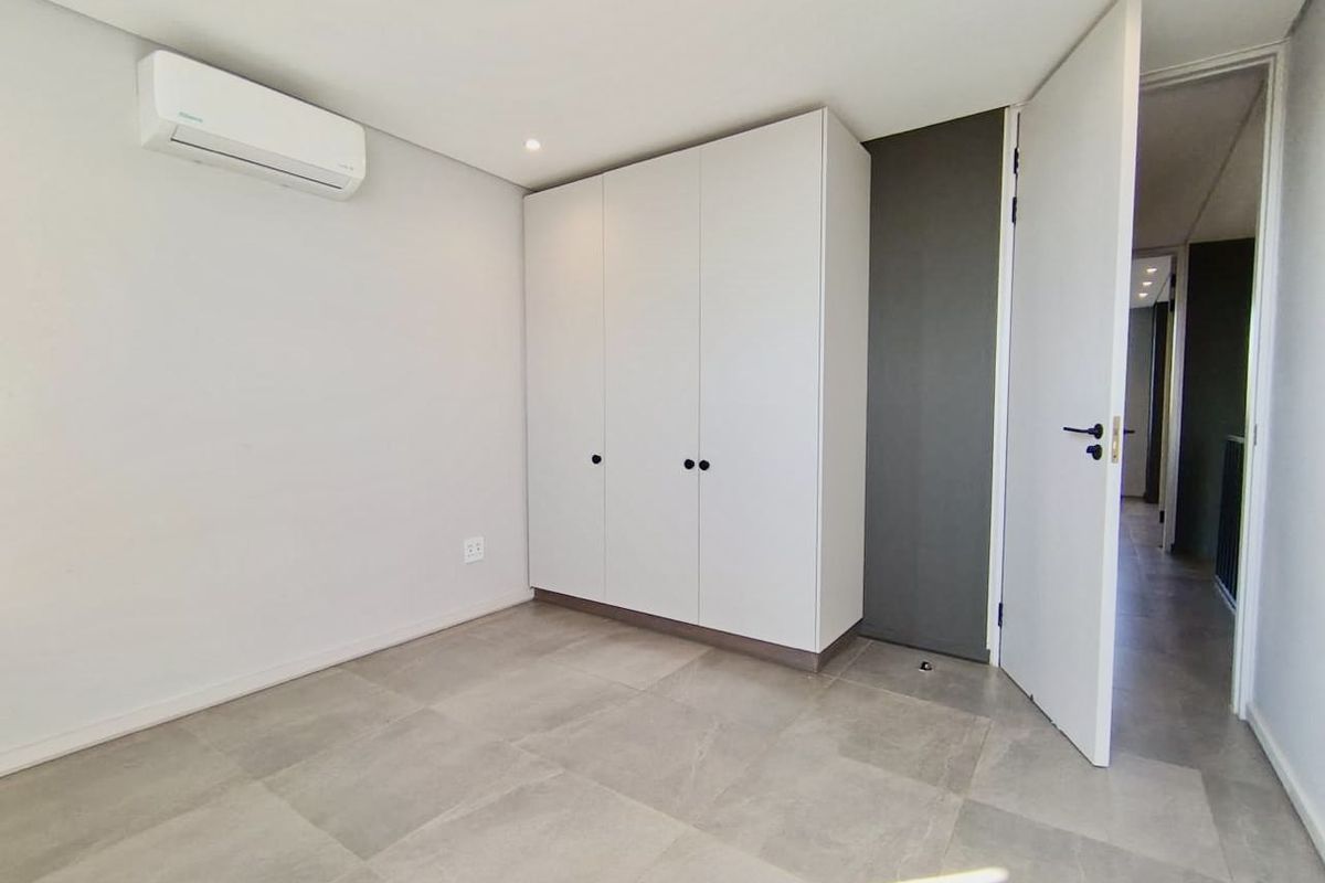 A bright, modern bedroom featuring neutral tiled flooring, built-in white cabinetry, and a wall-mounted air conditioning unit.