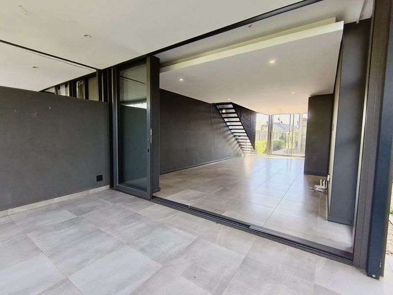 Spacious open-plan living area featuring large sliding glass doors, modern tiled flooring, and a sleek metal staircase leading to the upper level.