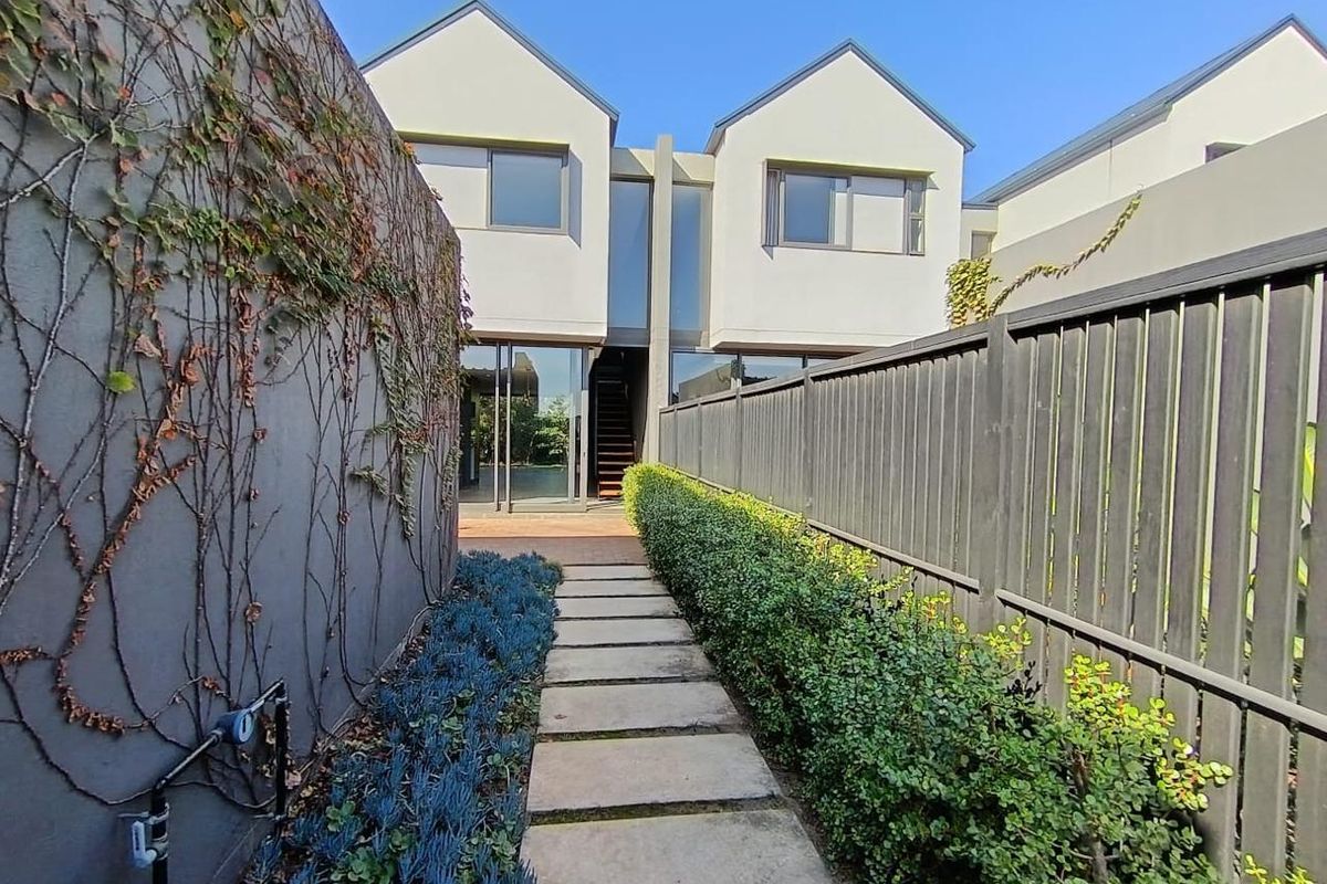 A rear view of the townhouse featuring a paved garden walkway, boundary fencing, and modern architectural facade with large glass sliding doors.