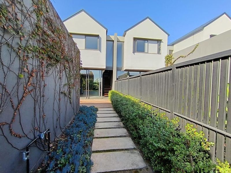 A rear view of the townhouse featuring a paved garden walkway, boundary fencing, and modern architectural facade with large glass sliding doors.