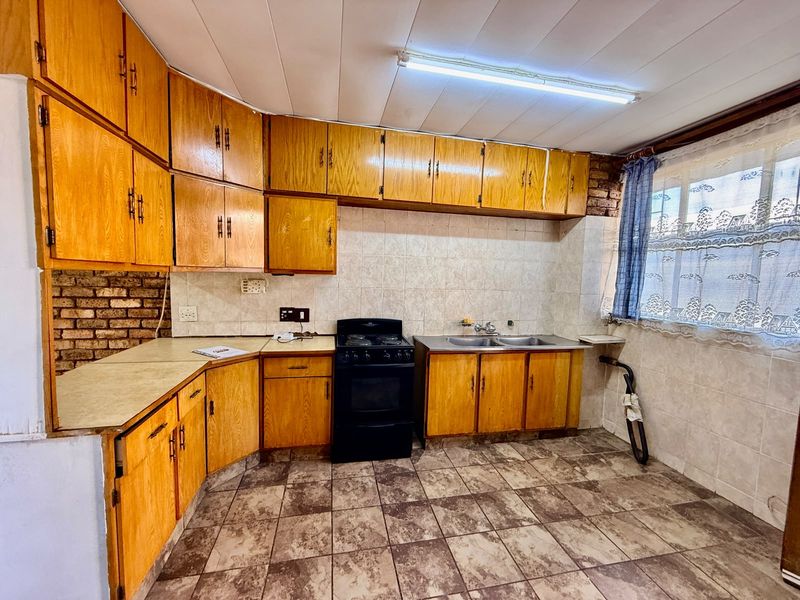 A dated kitchen featuring wooden cabinetry, laminate countertops, a double basin sink, and a freestanding stove, with tiled flooring and backsplash throughout.