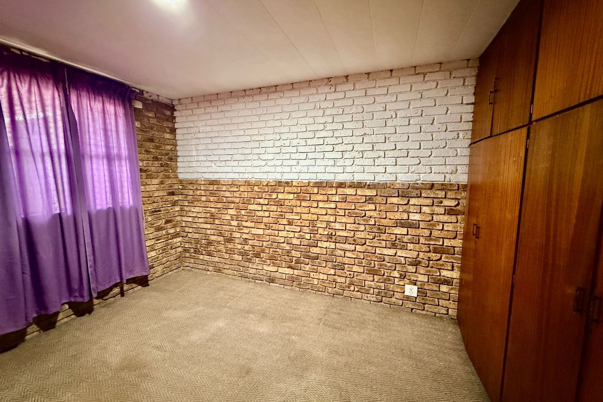 A bedroom featuring built-in wooden cupboards, carpeted flooring, and a distinctive half-painted exposed brick feature wall.
