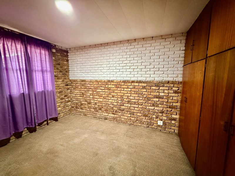 A bedroom featuring built-in wooden cupboards, carpeted flooring, and a distinctive half-painted exposed brick feature wall.