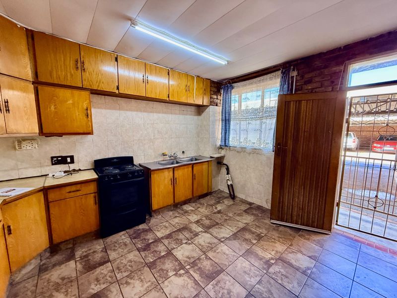 A dated kitchen featuring wooden cabinetry, tiled splashbacks, and a freestanding stove, with direct access to an exterior area through a security gate.