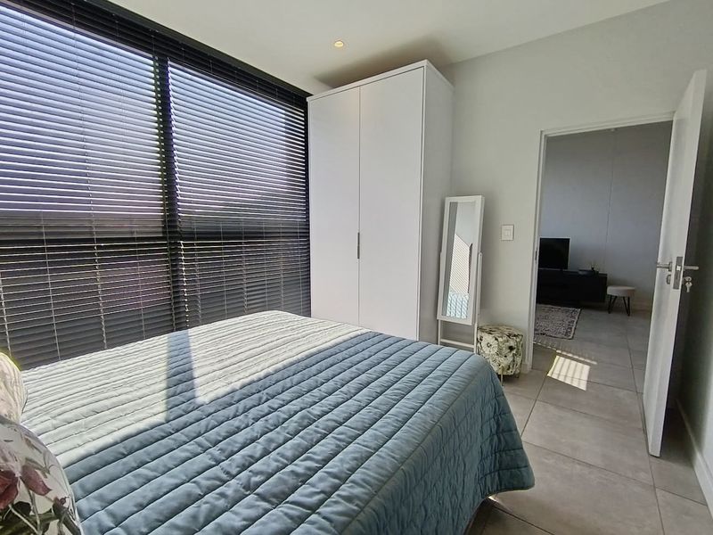 A bright bedroom featuring tiled flooring, a built-in wardrobe, and large windows fitted with venetian blinds, offering a clean and modern aesthetic.