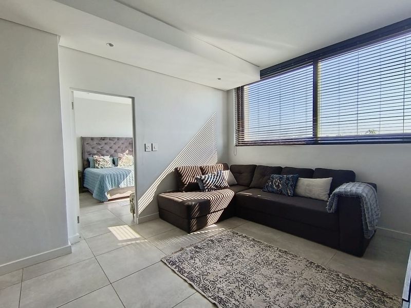 A bright living room featuring large windows with venetian blinds, tiled flooring, and recessed lighting, with an adjacent bedroom visible through the doorway.